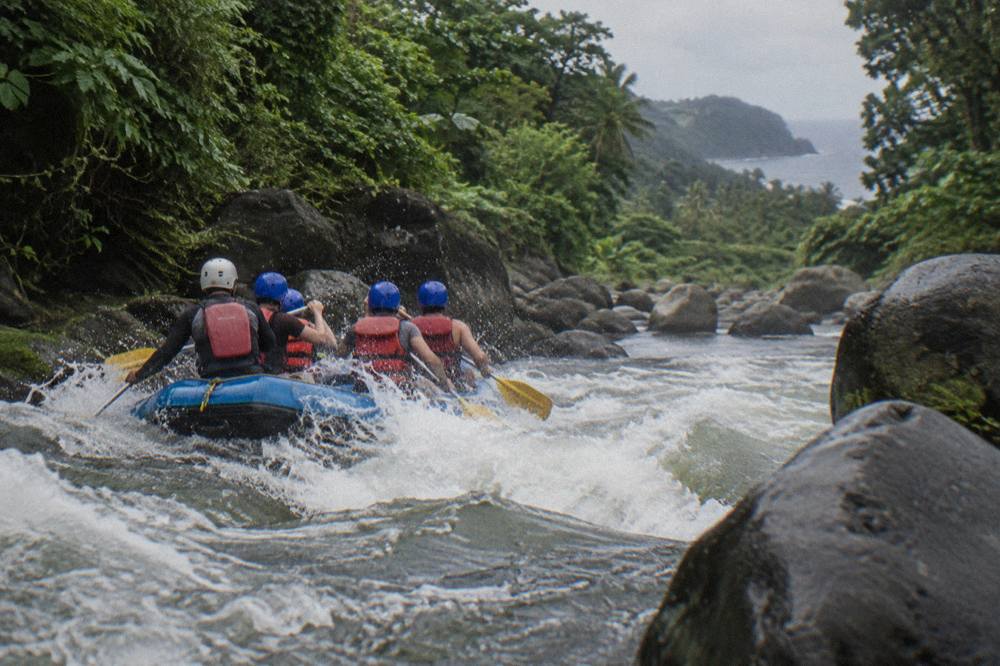 A raft with paddlers in helmets and life vests passing through a wave train in a narrow green river corridor