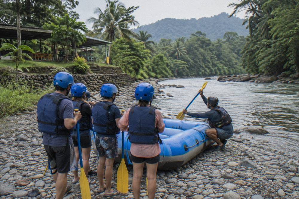 A guide explaining paddle commands to a family seated in an inflatable raft at a river put-in