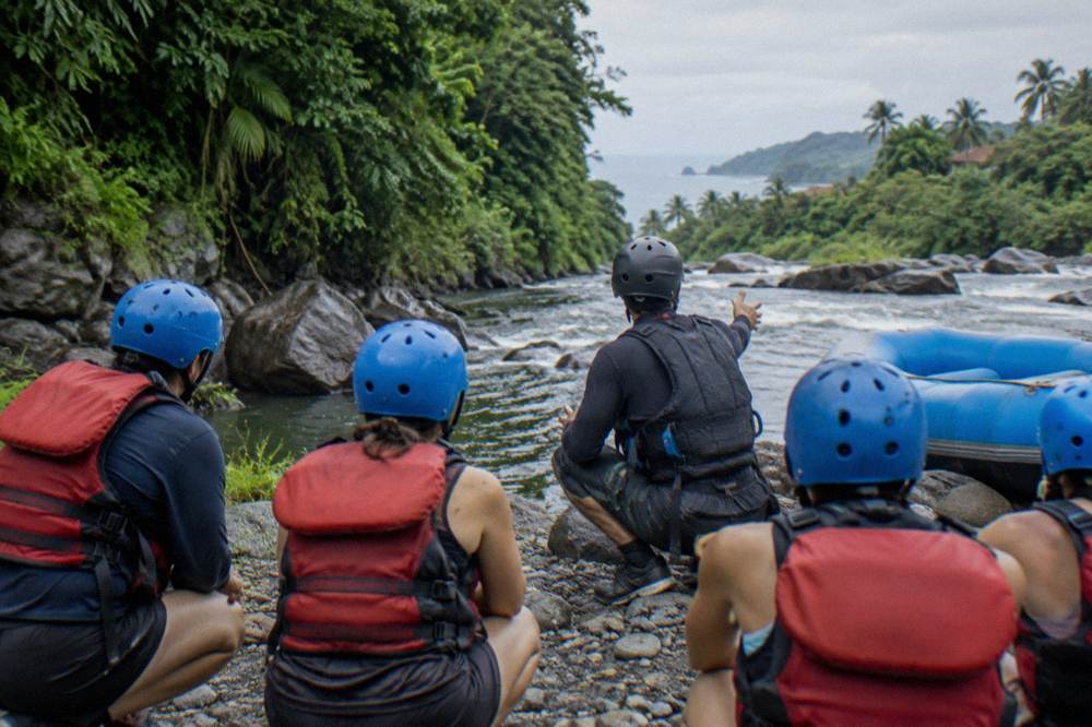 Rafters in helmets and life vests listening to a guide on a riverbank