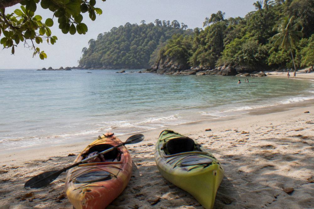 Kayaks resting on pale sand beside calm clear water with forested hills in the background