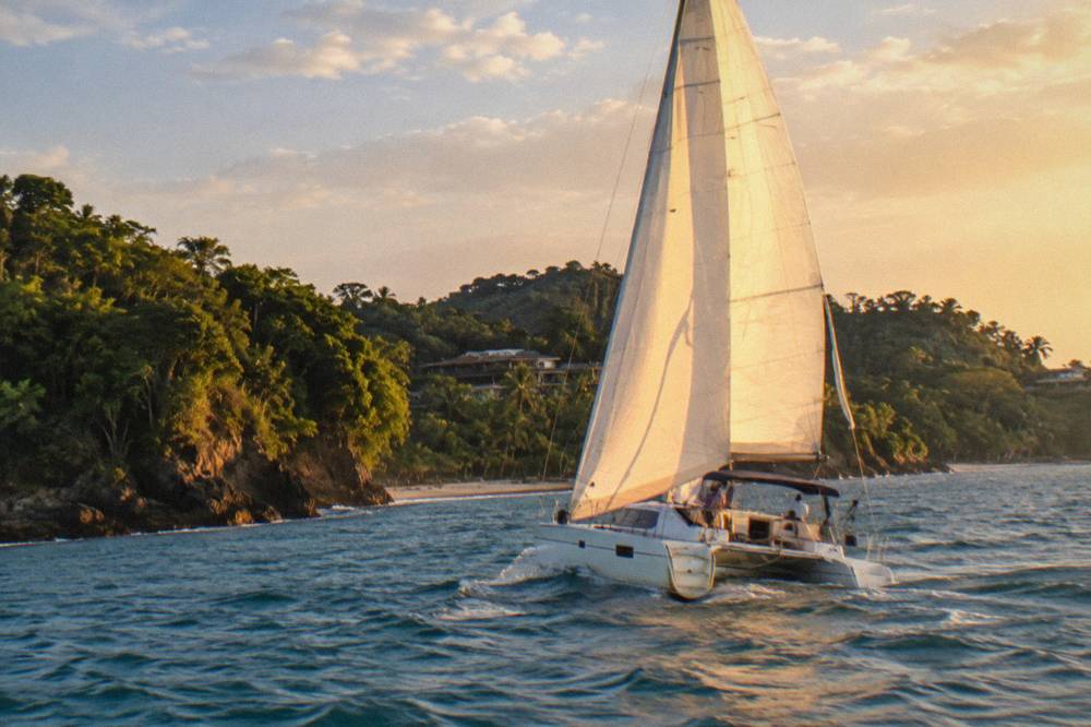 A sailing catamaran heeling under sail along a green coastline in late afternoon light