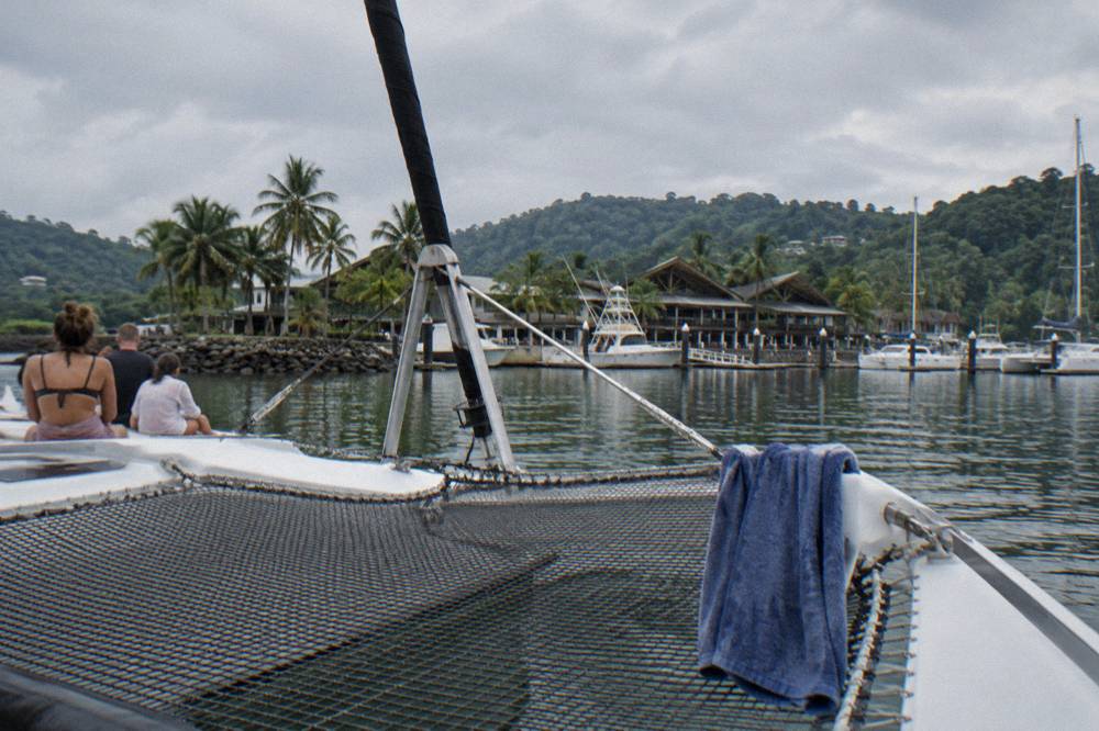 A wide catamaran deck with netting at the bow and passengers seated along the trampoline
