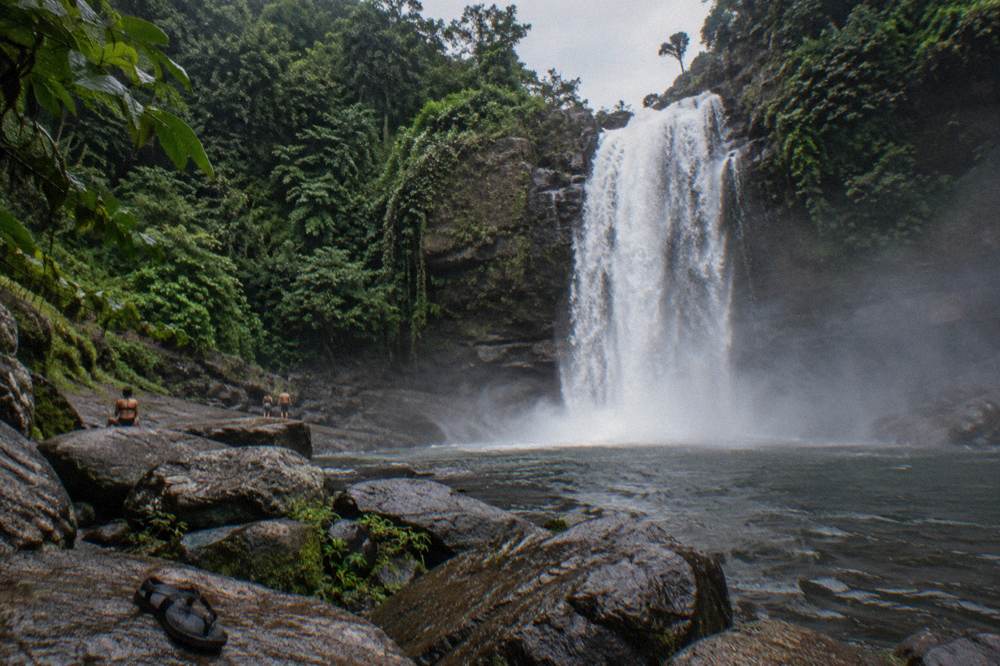 A tall waterfall dropping into a dark forest pool with rocks at its base
