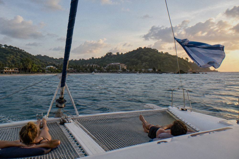 Open deck of a sailing catamaran in late afternoon light off a green coastline