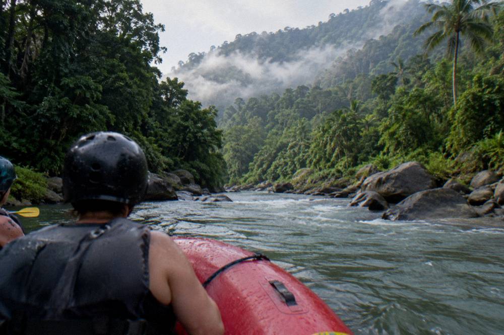 Raft paddling through a green rainforest river corridor with mist on the hills