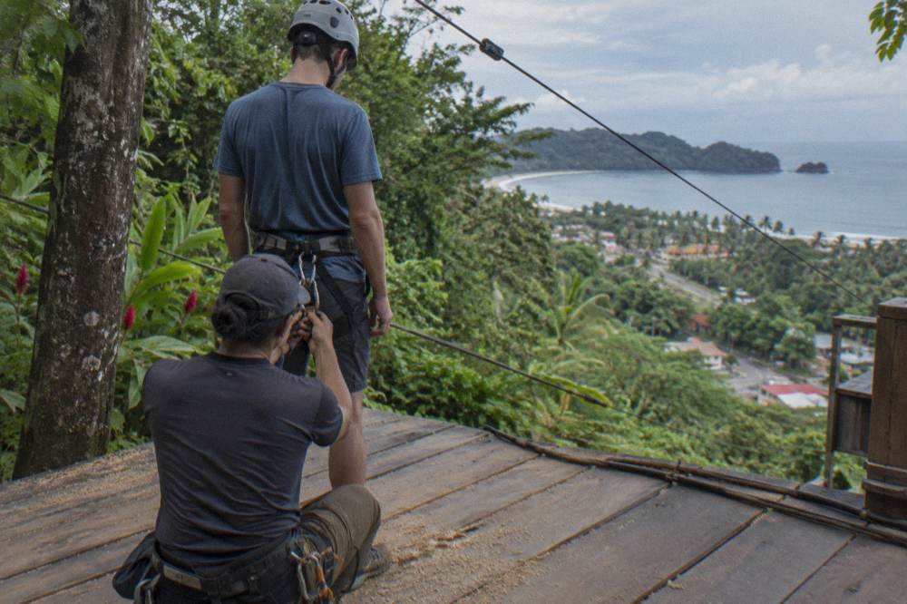 Guide checking a climber's harness and helmet on a wooden platform in the forest