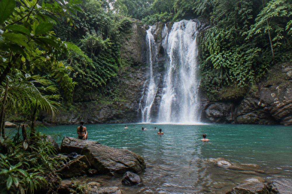 Turquoise pool at the base of a tall waterfall surrounded by dense green forest