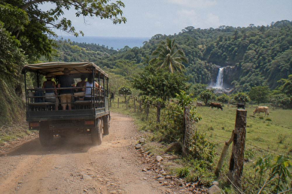 Open air truck carrying visitors along a dirt road through green hills
