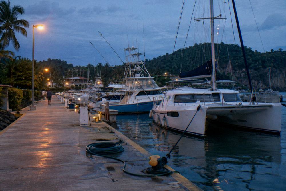 A marina dock at dusk with fishing boats and catamarans moored side by side