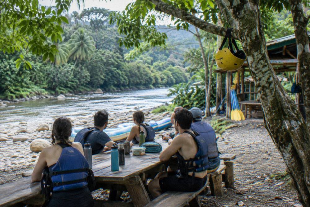 Rafting crew in helmets and vests eating lunch at a riverside table under trees