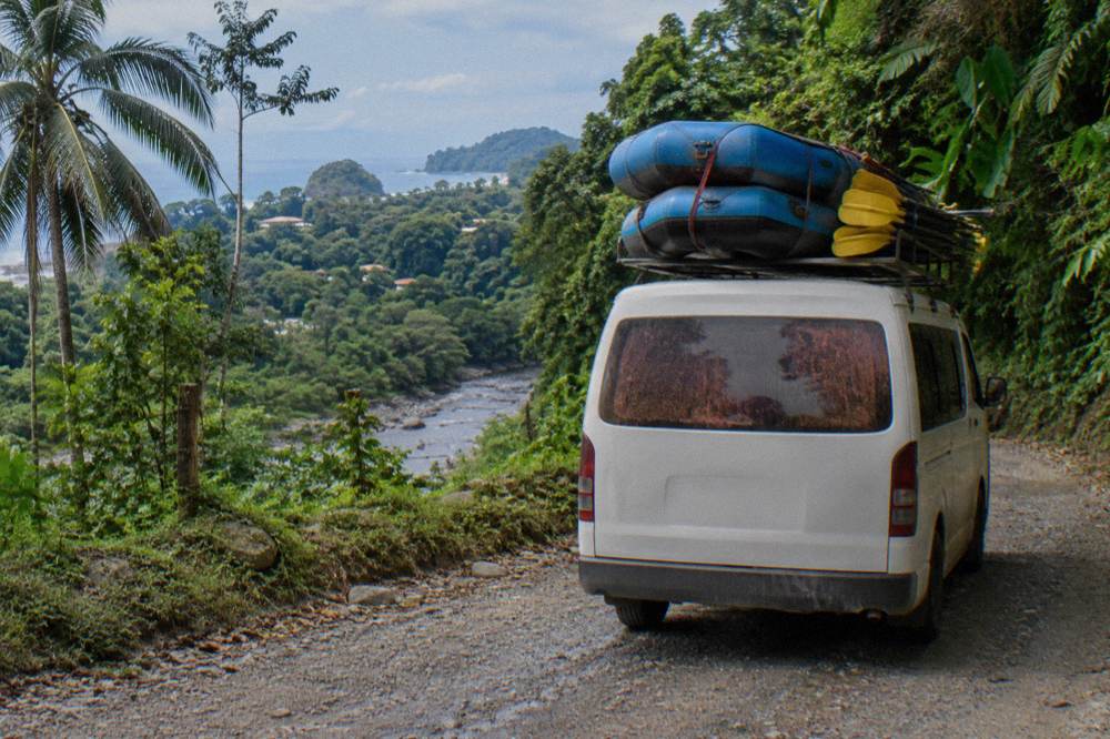 Van parked on a gravel road beside a jungle river with rafting equipment strapped to the roof