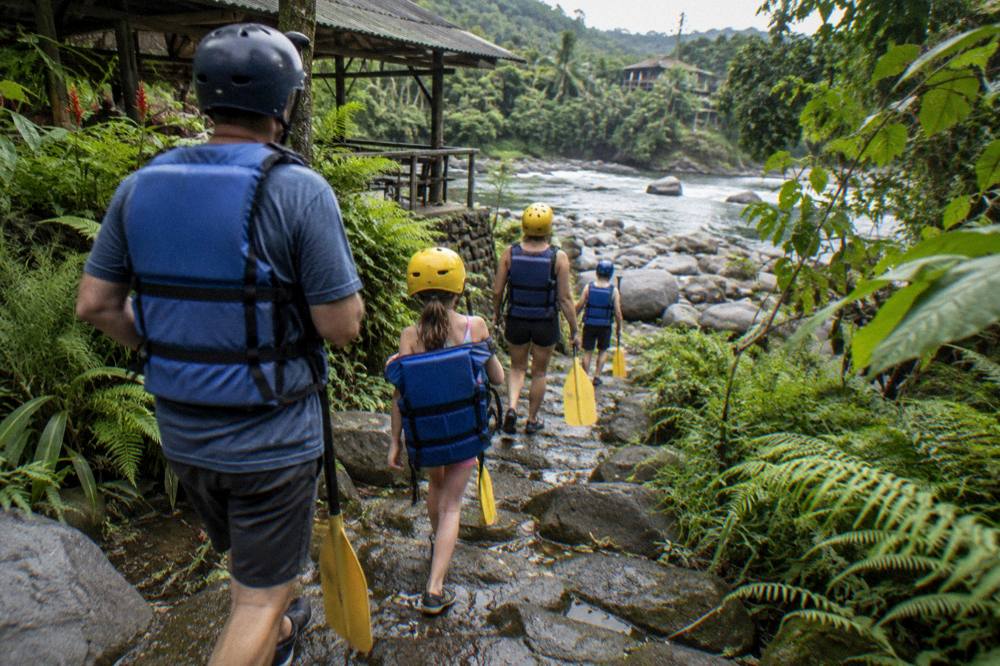 Family in helmets and life vests carrying paddles down a rocky path toward the river