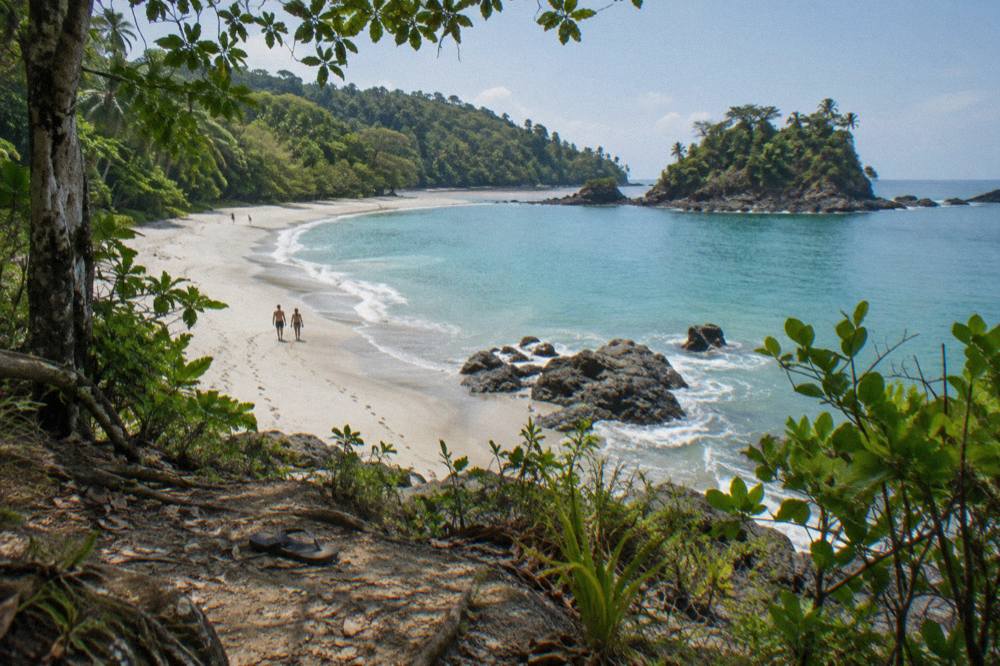 Wooded headland above a sandy beach with calm turquoise water and forested hills behind
