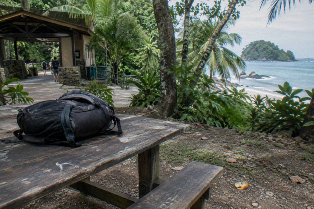 A shaded picnic table area beside a tropical forest edge with an empty backpack