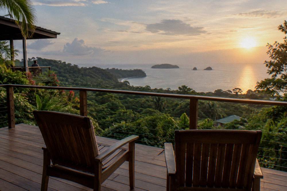 A hillside hotel terrace with two empty chairs looking over rainforest treetops to the Pacific